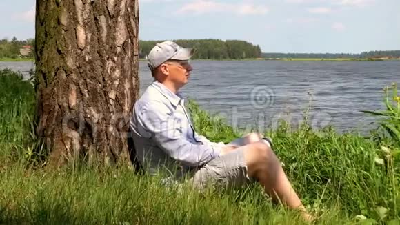 戴帽子的人坐在湖边的一棵树下喝水视频的预览图