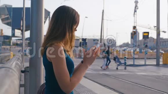 年轻迷人的女人坐在公共汽车或地铁车站女孩用她的手机输入信息在等她视频的预览图