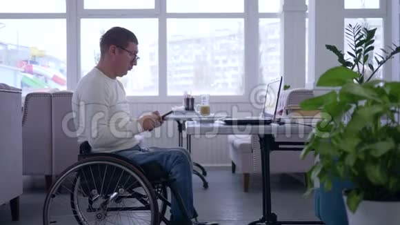 在线学习自由职业者残疾人在轮椅上戴眼镜使用手机与电脑坐在一起视频的预览图