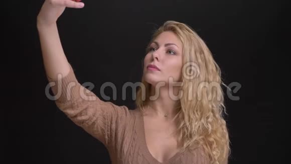 华丽的白种人长发女人在黑色背景下的智能手机上制作自拍照片的肖像视频的预览图