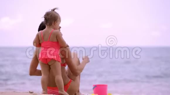 妈妈和女儿穿着粉红色的泳衣在一个美丽的海滩的岸边玩在沙滩上和孩子们海滩玩具妈妈视频的预览图