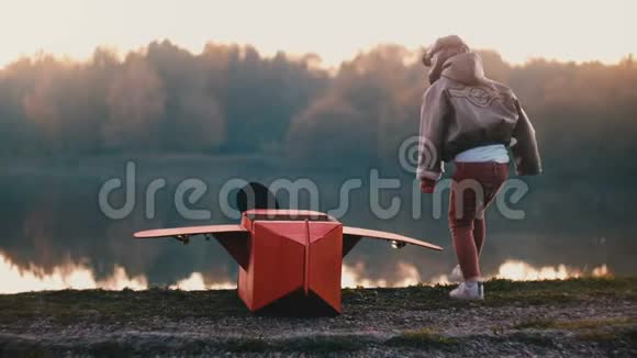 后景小男孩穿着古老的飞行员服装坐在惊人的森林湖与有趣的红色纸板飞机慢动作视频的预览图
