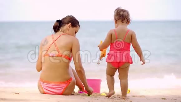 妈妈和女儿穿着粉红色的泳衣在一个美丽的海滩的岸边玩在沙滩上和孩子们海滩玩具视频的预览图