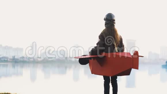 快乐的小飞行员女孩站在平静的湖城全景在纸板飞机上玩飞行员回首慢动作视频的预览图
