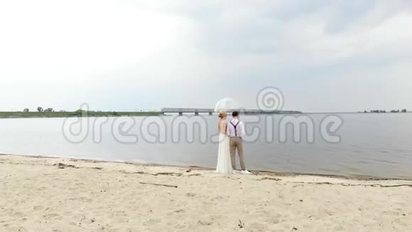 空中美丽的新婚夫妇站在海滩上在一把透明的伞下迎着蓝天河流和一大片视频的预览图