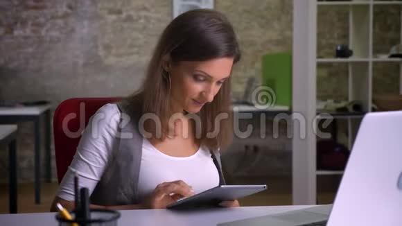 漂亮的女办公室工作人员在平板电脑上发短信在笔记本电脑前笑嘻嘻地坐着视频的预览图