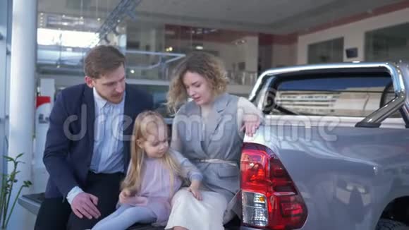 汽车销售中心年轻夫妇带着孩子选择汽车坐在后备箱里交流视频的预览图