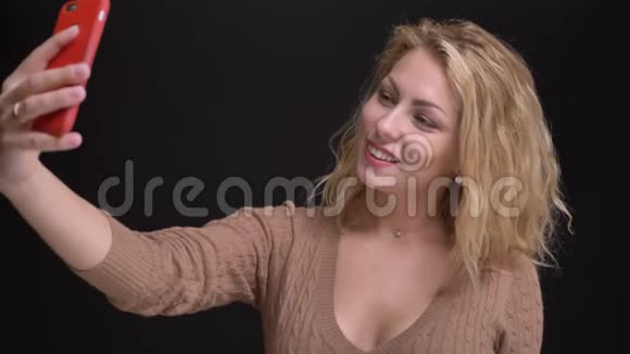 快乐的白种人长发女人微笑着在黑色背景下的智能手机上制作自拍照片的肖像视频的预览图