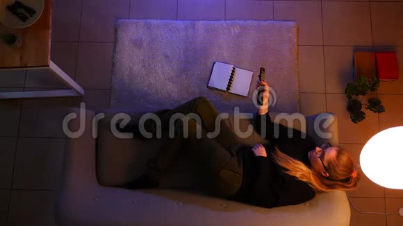 一位漂亮的女性躺在室内的沙发上看电视时在一间关灯的舒适公寓里拍摄特写镜头视频的预览图