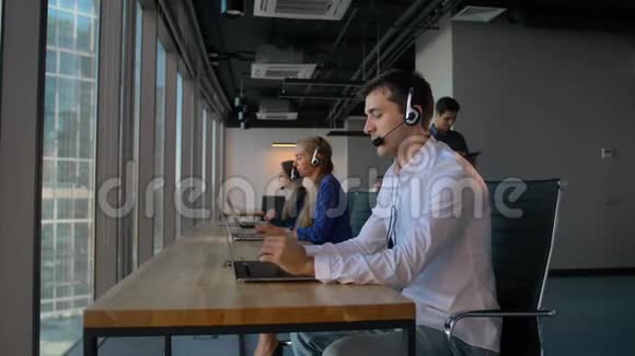 在呼叫中心办公室工作时穿着衬衫的男士使用带有笔记本电脑的耳机视频的预览图