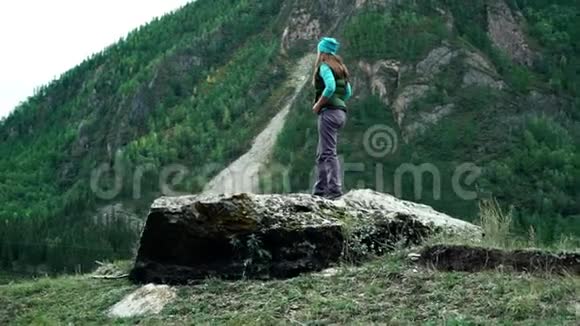 穿着背心和帽子的女旅行者站在树木覆盖的山上的一块大石头上视频的预览图