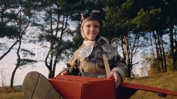 快乐的小飞行员男孩站在树下穿着纸板飞机服装戴着老式眼镜玩着飞行员慢动作视频的预览图