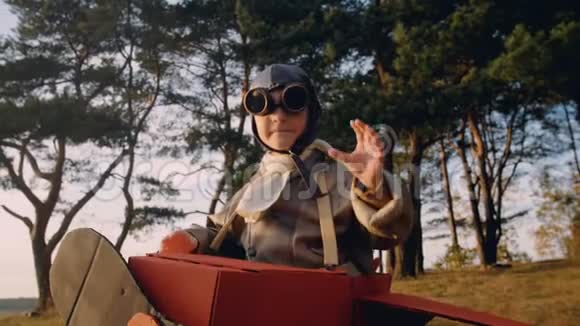 积极的小飞行员男孩在树下穿着有趣的纸板飞机服装戴着复古飞行员超级英雄眼镜慢镜头视频的预览图