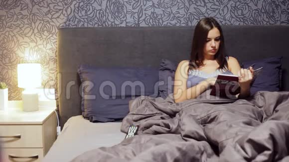 美丽的年轻女孩躺在卧室的床上读一本烹饪书晚上在家休息的女人视频的预览图