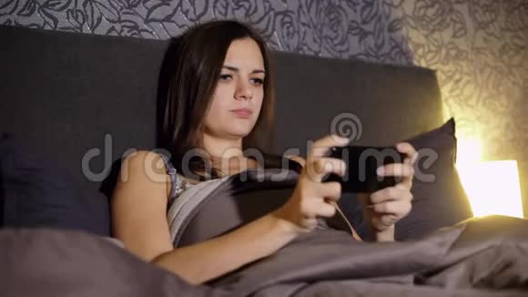 年轻女性在晚上躺在家里的床上玩智能手机游戏紧张的比赛输掉比赛视频的预览图