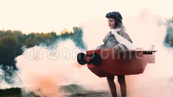 快乐的小飞行员男孩在有趣的纸板飞机服装与红色烟雾梦想飞行慢动作视频的预览图