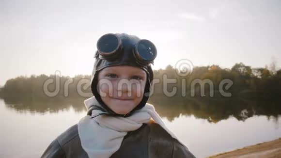 快乐的小飞行员男孩戴着复古的飞行员眼镜沿着日落湖奔跑假装是一架飞机慢动作视频的预览图