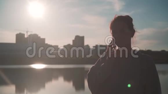 一个年轻的女孩在日落时分的阳光下在城市背景下的湖上用手机说话视频的预览图