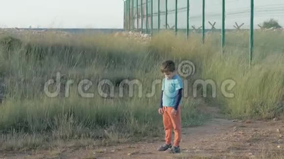 穿着脏衣服的小男孩孤独地站在靠近金属栅栏的田野里看着障碍物感觉情感避难所的概念视频的预览图