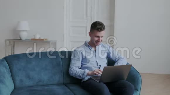 年轻快乐的时髦男人穿着蓝色衬衫和他的朋友聊天他微笑着用短信在笔记本电脑上打字视频的预览图