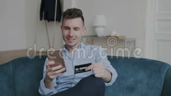 网上购物的迷人年轻人在家里的沙发上使用电话和信用卡他想象他的购买视频的预览图