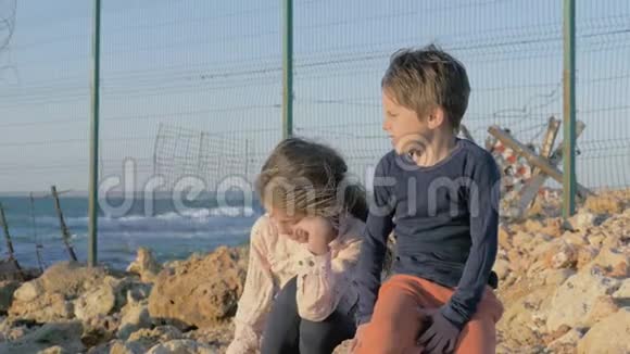 两个可怜的难民儿童男孩女孩坐在海边的石头上用铁丝网把这个州隔开视频的预览图