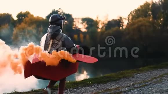 镜头跟随快乐的小飞行员男孩沿着日落湖在有趣的纸板飞机与彩色烟雾慢动作视频的预览图