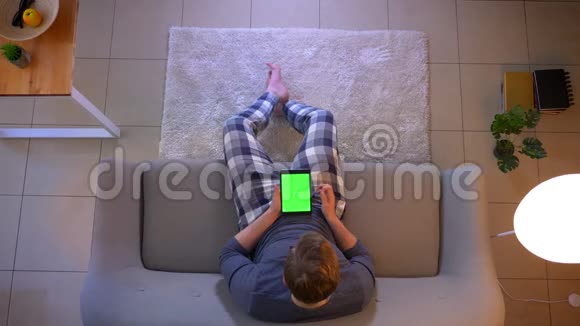 在舒适的室内沙发上坐着绿色屏幕的平板电脑上一位穿着随意的年轻男性的特写镜头视频的预览图