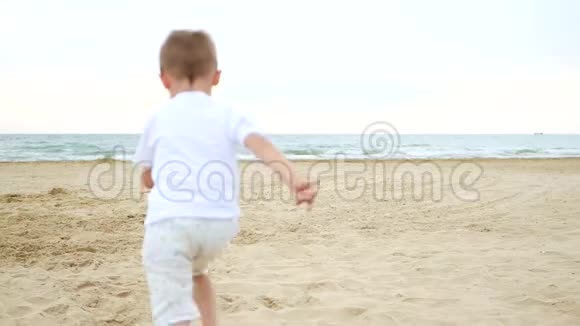 摄像机的焦点在奔跑的孩子后面平稳地移动小快乐男孩沙滩到海边小孩玩耍视频的预览图