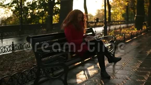 年轻的女士正在看书红头可爱的女孩正坐在公园的长椅上秋天五彩缤纷的景色镜头耀斑放松一下视频的预览图