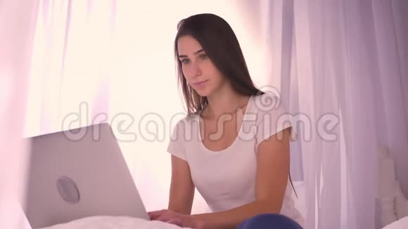 年轻迷人的少女在笔记本电脑上打字看着坐在家里床上的照相机的特写照片视频的预览图
