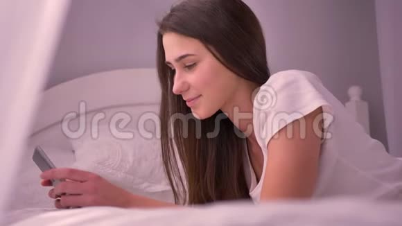一位年轻的白种人女性在床上躺着然后看着摄像机微笑着用平板电脑拍摄她的特写照片视频的预览图