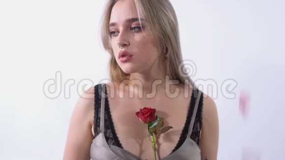 在一个明亮的工作室里一个悲伤的女人站在一个白色背景上鼻子被刺穿玫瑰花瓣从上面倾泻而下视频的预览图