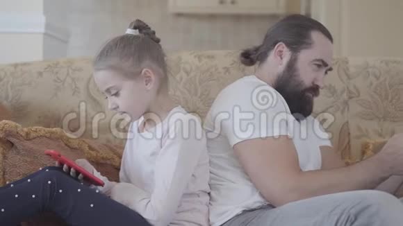 可爱的女儿和她的父亲坐在客厅的沙发上带着智能手机在舒适的日子里不寒而栗父亲和视频的预览图