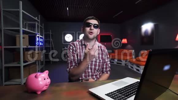 有魅力的年轻人带着太阳镜对着镜头说话有猪和笔记本电脑在桌子上视频的预览图