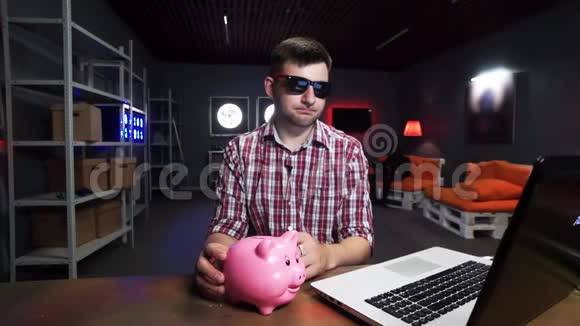 年轻的大胡子男性坐在桌子后面手提电脑拿着塑料猪说话视频的预览图