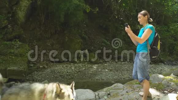 骑着漂亮的女人背着背包用智能手机在山河畔与马拉木特狗在丛林中合影视频的预览图