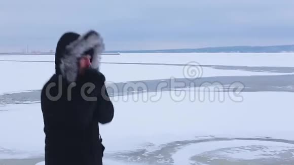 身穿黑色夹克衫头戴毛皮帽的年轻人站在结冰的河底从寒冷中跳下来试图保暖视频的预览图