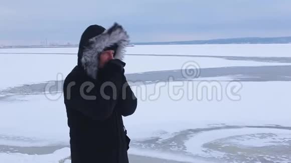 身穿黑色夹克衫头戴毛皮帽的年轻人站在结冰的河底双手戴手套取暖视频的预览图