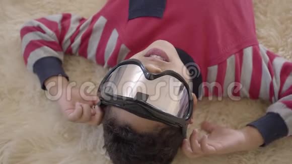 美国黑人小男孩躺在米黄色蓬松地毯上的肖像眼睛上戴着彩弹眼镜视频的预览图