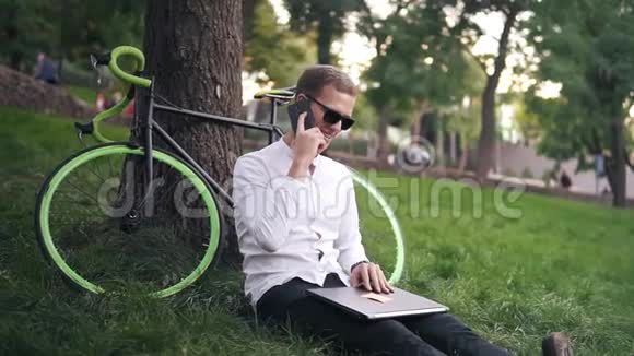 年轻的成功商人穿着白色衬衫和黑色眼镜男人坐在草地上手提电脑继续说话视频的预览图
