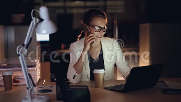 可爱的女孩公司经理用手机说话用笔记本电脑坐在黑暗的办公室里微笑着视频的预览图