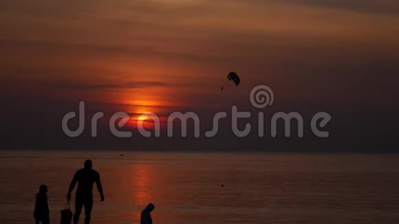 日落时在热带海滩进行降落伞活动人们在海滩上散步和在海里游泳的剪影冷静视频的预览图
