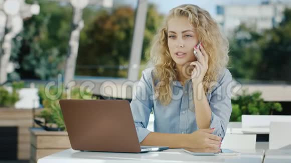 很有魅力的商务女性在打电话坐在露台上的咖啡馆里拿着笔记本电脑视频的预览图