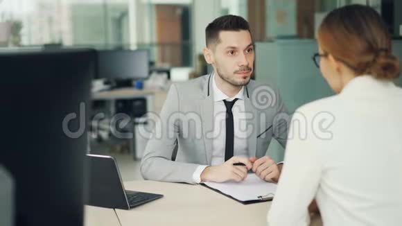 一位开朗的男性面试官正在和一位女性求职者交谈她坐在现代办公室的办公桌旁微笑着视频的预览图