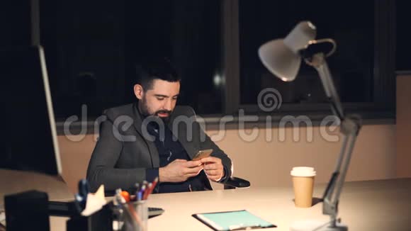 一个英俊的留着胡子的年轻人正在使用智能手机触摸屏幕和微笑独自在办公室工作到深夜视频的预览图