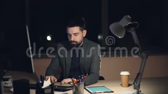 一个穿着西装打着领带的疲惫的年轻人正在办公室工作晚上坐在黑暗的房间里的办公桌前看着视频的预览图