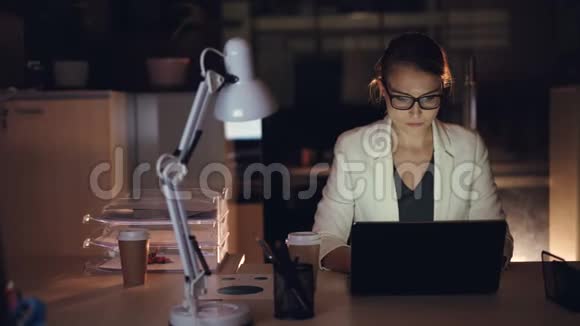 穿着正式服装的漂亮女孩独自在笔记本电脑上工作在黑暗的办公室深夜完成紧急工作视频的预览图