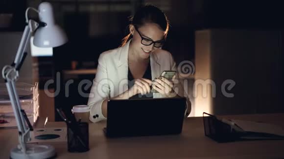 漂亮的金发女郎正在使用智能手机发短信深夜独自坐在办公室里微笑现代小玩意硬的视频的预览图
