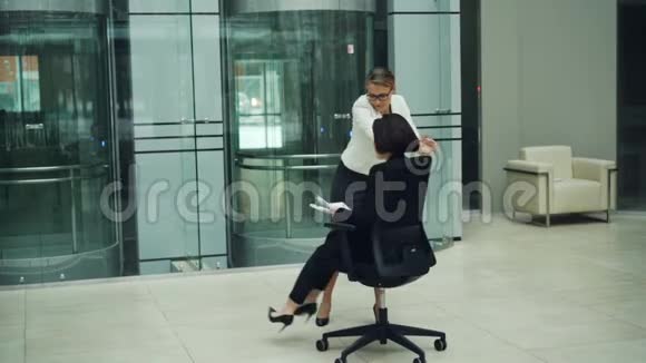 快乐的年轻女性在办公室大堂享受乐趣的慢动作坐在摇椅上旋转并投掷视频的预览图